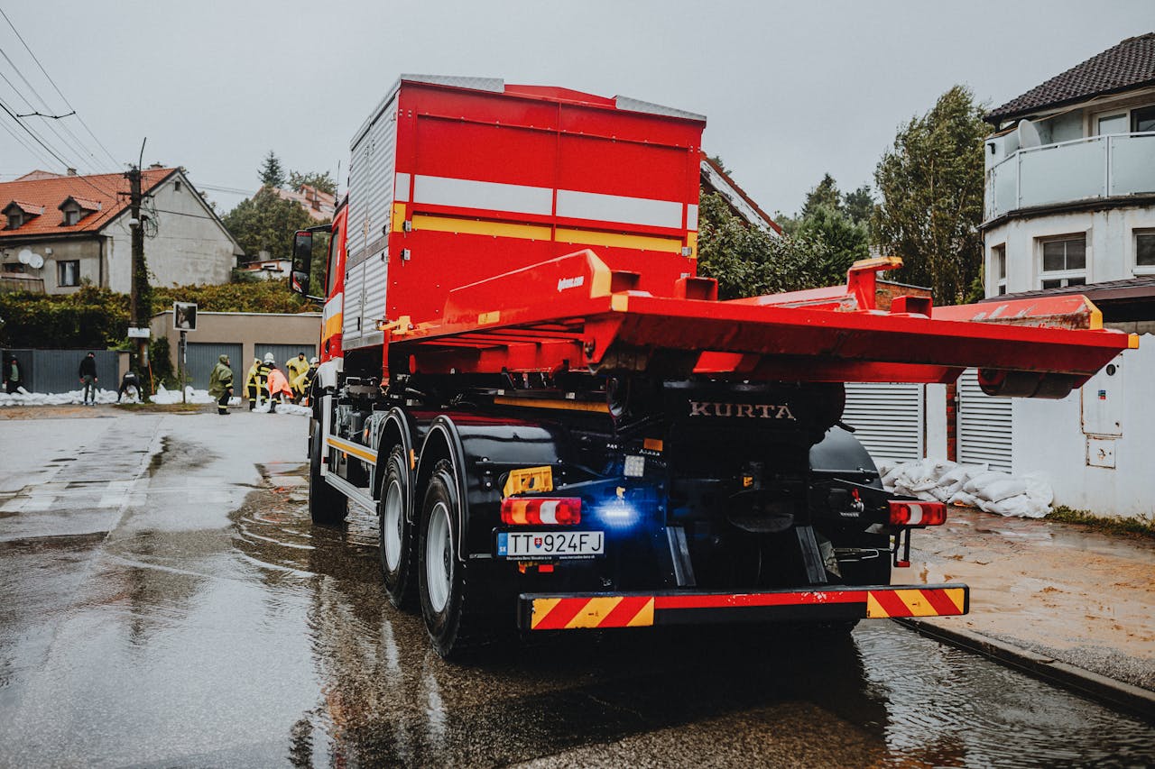 A red tow truck parked on a wet street in an urban neighborhood with workers in the background.